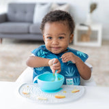 A curly-haired toddler smiles, enjoying a meal from a blue bowl on a Bumkins Silicone Sensory Placemat: Marble while wearing a blue bib.