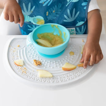A child enjoys applesauce and apple slices in a blue bowl on Bumkins Silicone Sensory Placemat: Marble at a white table.
