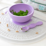 A purple baby bowl with mashed peas and a spoon sits on a Bumkins Silicone Sensory Placemat: Marble.