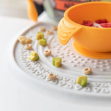 Close up of an orange bowl on a Bumkins Silicone Sensory Placemat: Marble with scattered cereal and avocado pieces.