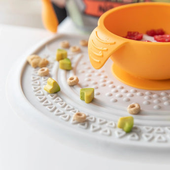 Close up of an orange bowl on a Bumkins Silicone Sensory Placemat: Marble with scattered cereal and avocado pieces.