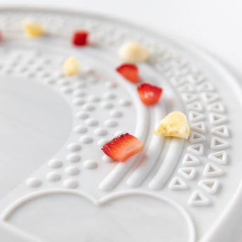 Close-up of Bumkins Silicone Sensory Placemat: Marble strawberry and banana pieces scattered on top.