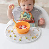 A baby in a colorful bib enjoys cereal and fruits from an orange bowl on a Bumkins Silicone Sensory Placemat: Marble.