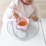 A toddler enjoys applesauce from a pink bowl on a Bumkins Silicone Sensory Placemat: Marble at a white table.