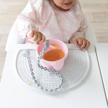 A toddler enjoys applesauce from a pink bowl on a Bumkins Silicone Sensory Placemat: Marble at a white table.