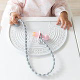 A toddler in high chair with a pink utensil on a silicone tether attached to a Bumkins Silicone Sensory Placemat: Marble on a white table.