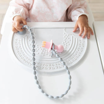 A toddler in high chair with a pink utensil on a silicone tether attached to a Bumkins Silicone Sensory Placemat: Marble on a white table.