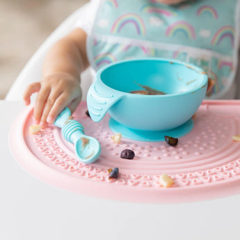 A baby in a high chair holds a blue spoon next to a blue bowl on top of a Bumkins Silicone Sensory Placemat: Pink.