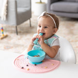 A baby with a headband smiles in a high chair, biting a spoon while a bowl rests on a Bumkins Silicone Sensory Placemat: Pink.