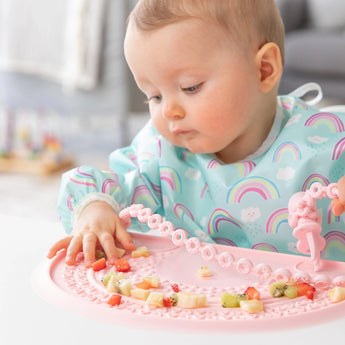 A baby plays with food on a Bumkins Silicone Sensory Placemat: Pink while wearing a rainbow patterned bib.