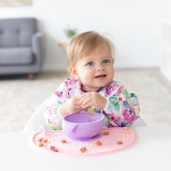 A toddler wearing a colorful bib smiles next to a purple bowl on a Bumkins Silicone Sensory Placemat: Pink on a white table.