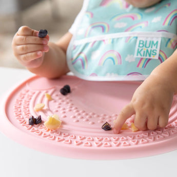 Close-up of a child playing with pieces of food on a Bumkins Silicone Sensory Placemat: Pink at a white table.