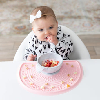 A baby in a heart-patterned bib with a white bow, eats from a gray bowl on a Bumkins Silicone Sensory Placemat: Pink on a white table.