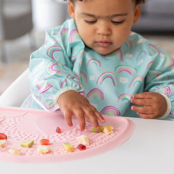 A toddler in a rainbow bib explores food on a Bumkins Silicone Sensory Placemat: Pink at a white table.