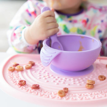Close-up of a child eating from a purple bowl with a spoon on a Bumkins Silicone Sensory Placemat: Pink with scattered cereal pieces.