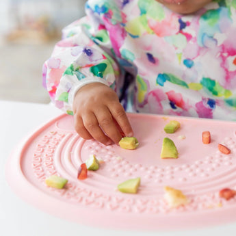 Close-up of a child in a colorful bib reaching for avocado on a vibrant Bumkins Silicone Sensory Placemat: Pink.