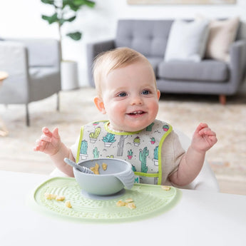 A smiling baby in a cactus bib sits at a white table, with a gray bowl on Bumkins Silicone Sensory Placemat: Sage. 