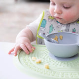 A baby explores food on a Bumkins Silicone Sensory Placemat: Sage with a nearby gray bowl and spoon, wearing a cactus-patterned bib.