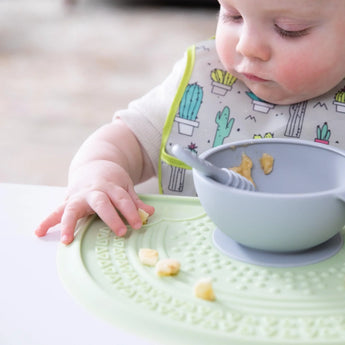 A baby explores food on a Bumkins Silicone Sensory Placemat: Sage with a nearby gray bowl and spoon, wearing a cactus-patterned bib.