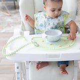 A baby in a high chair plays with cereal on a Bumkins Silicone Sensory Placemat: Sage with a gray bowl on top, wearing a cactus bib.