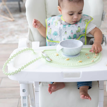 A baby in a high chair plays with cereal on a Bumkins Silicone Sensory Placemat: Sage with a gray bowl on top, wearing a cactus bib.