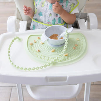 A baby in a high chair enjoys cereal from a bowl on a Bumkins Silicone Sensory Placemat: Sage.