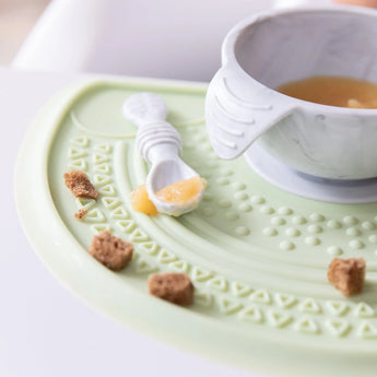A gray bowl and spoon sits on top of a Bumkins Silicone Sensory Placemat: Sage scattered with food on a high chair tray.