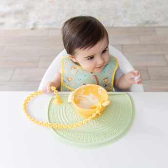 A baby in high chair, wearing a patterned bib, plays with yogurt in a bowl on a Bumkins Silicone Sensory Placemat: Sage.