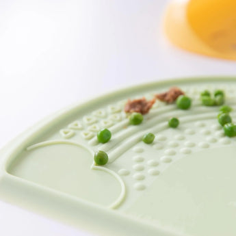 Close-up of Bumkins Silicone Sensory Placemat: Sage on a white background, with peas scattered on top.