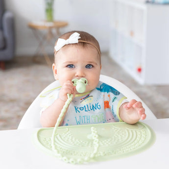 A baby chews on a silicone baby utensil, with a Bumkins Silicone Sensory Placemat: Sage on a white table nearby.