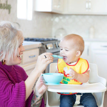 Adult with short blonde hair feeds a smiling toddler in a high chair using a blue spoon and bowl; child wears Bumkins Super Mario™ SuperBib.