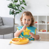 A young child in the living room sports a Bumkins SuperBib® from the Super Mario™ Expressions pack, using a yellow plate near plants and shelves.