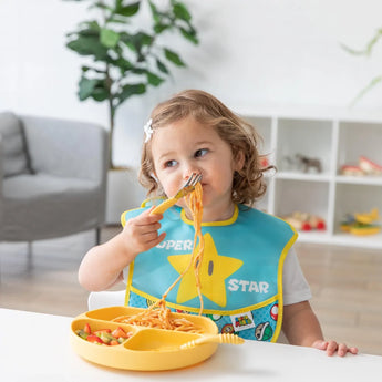 Toddler enjoying spaghetti from a yellow plate, wearing a Super Mario™ Expressions SuperBib® by Bumkins, with a plant nearby.