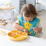 A toddler enjoys spaghetti and veggies in a SuperBib® by Bumkins at a white table, using a yellow plate.