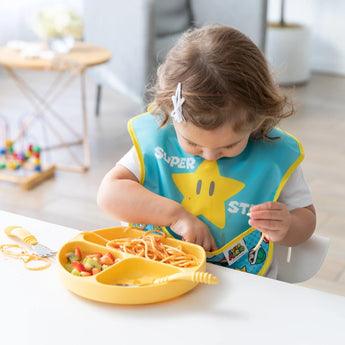 A toddler enjoys spaghetti and veggies in a SuperBib® by Bumkins at a white table, using a yellow plate.