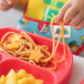 Childs hand holds spaghetti on red plate with star cheese, pineapple side, and Bumkins Super Mario™ SuperBib® 2 Pack—stay mess-free!.