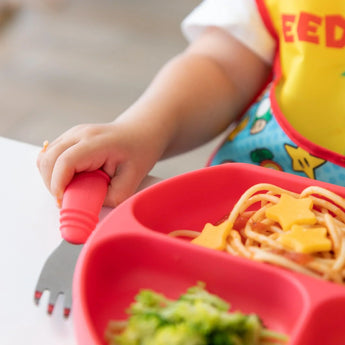 A child holds a fork by a divided plate with spaghetti, cheese, and broccoli, wearing a Bumkins Super Mario™ Expressions SuperBib®.