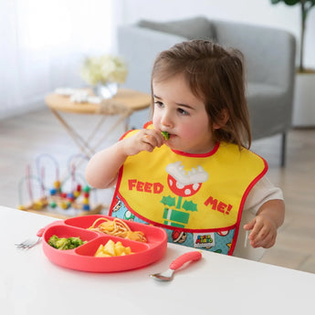A toddler in a Bumkins Super Mario SuperBib enjoys broccoli, pineapple, and spaghetti from a sectioned plate at the table.