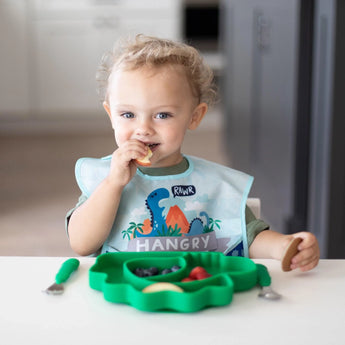 A young child enjoys snacks with a Bumkins dinosaur-themed SuperBib® with crumb catcher and a green dino footprint-shaped plate.