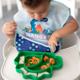 A child in a high chair uses a dinosaur SuperBib and eats from a dinosaur-shaped plate featuring Bumkins SuperBib®: Dinosaurs design.