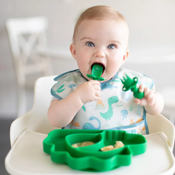 A baby in a high chair uses a green dinosaur-shaped spoon, matching plate, and wears a Bumkins SuperBib® with dinosaur prints.