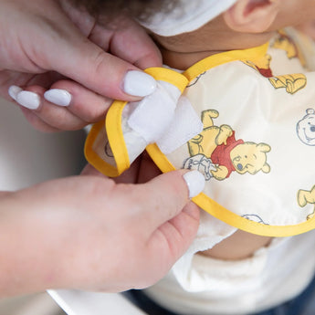 Close-up of adult hands adjusting the hook-and-loop closure of a Bumkins SuperBib® 3 Pack: Pooh Bear and Friends around a child's neck.