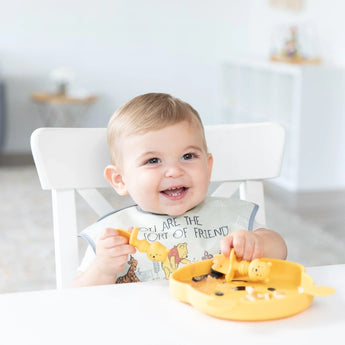A baby in a chair holds a yellow spoon and plate, wearing a Bumkins SuperBib® 3 Pack: Pooh Bear and Friends in a bright room.