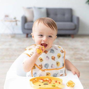 A baby in a high chair wearing a yellow Bumkins SuperBib® 3 Pack: Pooh Bear and Friends eats with a spoon; a gray sofa is in the background.