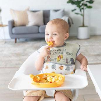 A baby in a Bumkins SuperBib® 3 Pack: Pooh Bear and Friends sits in a high chair, eating from a yellow plate in a living room.