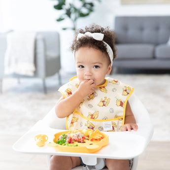 A toddler in a yellow Bumkins SuperBib® 3 Pack: Pooh Bear and Friends sits in a high chair, enjoying food from a yellow plate.
