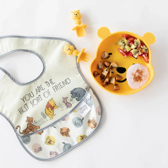A Bumkins SuperBib® 3 Pack: Pooh Bear and Friends lays flat on a white background next to a bear-shaped plate of food and matching utensils.