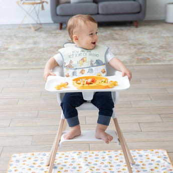A baby in high chair beams, wearing a Bumkins SuperBib® 3 Pack: Pooh Bear and Friends with a yellow plate of food on the tray.