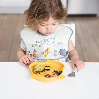 A child wears a Bumkins SuperBib® 3 Pack: Pooh Bear and Friends while eating from a yellow bear-shaped plate at a white table.
