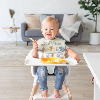 A toddler wearing a Bumkins SuperBib® 3 Pack: Pooh Bear and Friends sits in a high chair with a plate of food and a living room backdrop.